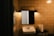 A dimly lit bathroom scene with beige tiled walls. A rectangular mirror is mounted above a white ceramic sink. There are two wall-mounted lights above the mirror, casting a warm glow. A metal stand holds a roll of paper towels and a glass.