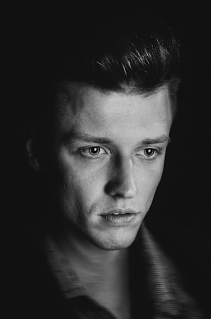 A monochrome portrait of a young man with short, neatly styled hair, gazing slightly downward. His face is illuminated softly, emphasizing his focused expression and detailed features. The background is dark, drawing attention to his contemplative demeanor.