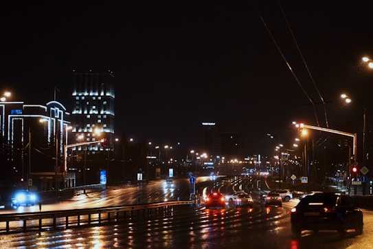 A nighttime cityscape featuring a wet road reflecting streetlights and headlights from passing cars. Tall buildings in the background are illuminated, adding a sleek urban feel. The sky is dark, with multiple streetlights lining the road and a few vehicles visible.