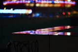 A dark urban scene with vibrant neon lights reflecting off a shiny surface. The defocused background features circular and linear patterns in vivid red, blue, and yellow colors, suggesting a lively city atmosphere.