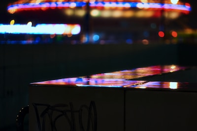 A dark urban scene with vibrant neon lights reflecting off a shiny surface. The defocused background features circular and linear patterns in vivid red, blue, and yellow colors, suggesting a lively city atmosphere.