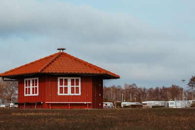A small, red wooden building with a tiled roof is situated in an open field. The structure features white-framed windows and is surrounded by a grassy area. In the background, there are bare trees and several trailers or prefabricated houses. The sky is mostly overcast with some patches of blue.