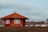 A small, red wooden building with a tiled roof is situated in an open field. The structure features white-framed windows and is surrounded by a grassy area. In the background, there are bare trees and several trailers or prefabricated houses. The sky is mostly overcast with some patches of blue.