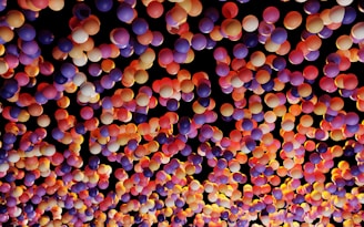 A vibrant and colorful array of balloons with various shades of purple, pink, orange, and yellow against a dark background, creating a festive and lively atmosphere.