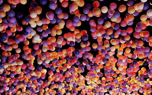 A vibrant and colorful array of balloons with various shades of purple, pink, orange, and yellow against a dark background, creating a festive and lively atmosphere.