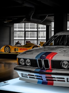 A showroom setting displays two racecars. In the foreground, a BMW with classic racing stripes in blue, red, and purple is prominently featured. In the background, a yellow racecar with sponsor logos, including DHl and Continental, is visible.