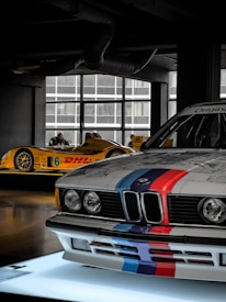 A showroom setting displays two racecars. In the foreground, a BMW with classic racing stripes in blue, red, and purple is prominently featured. In the background, a yellow racecar with sponsor logos, including DHl and Continental, is visible.