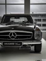 A vintage car with a shiny black exterior is displayed indoors, with the view of an industrial-looking building through large windows. The car features a prominent front grille adorned with a silver emblem and a classic design style.
