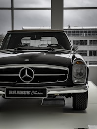 A vintage car with a shiny black exterior is displayed indoors, with the view of an industrial-looking building through large windows. The car features a prominent front grille adorned with a silver emblem and a classic design style.