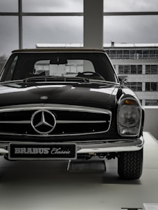 A vintage car with a shiny black exterior is displayed indoors, with the view of an industrial-looking building through large windows. The car features a prominent front grille adorned with a silver emblem and a classic design style.