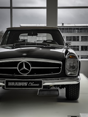 A vintage car with a shiny black exterior is displayed indoors, with the view of an industrial-looking building through large windows. The car features a prominent front grille adorned with a silver emblem and a classic design style.