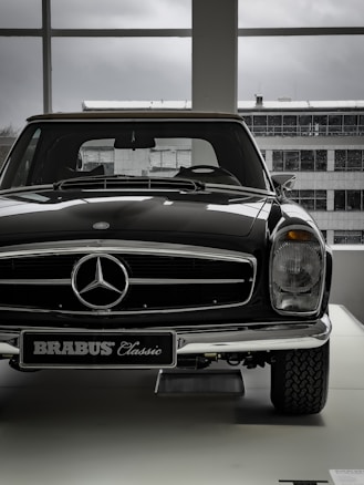 A vintage car with a shiny black exterior is displayed indoors, with the view of an industrial-looking building through large windows. The car features a prominent front grille adorned with a silver emblem and a classic design style.