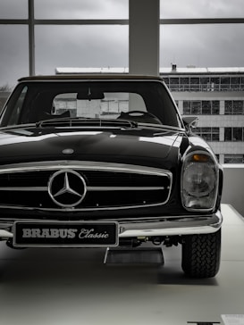 A vintage car with a shiny black exterior is displayed indoors, with the view of an industrial-looking building through large windows. The car features a prominent front grille adorned with a silver emblem and a classic design style.