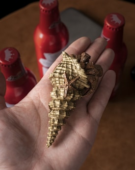 A hand holding a detailed seashell sculpture with a small starfish attached to it. In the background, there are three red bottles, possibly beer bottles, standing upright on a brown surface.