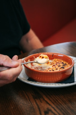 A person holds a spoon above an orange ramekin filled with a creamy dish, likely crème brûlée, placed on a white plate. The food has a caramelized top and is being scooped gently.