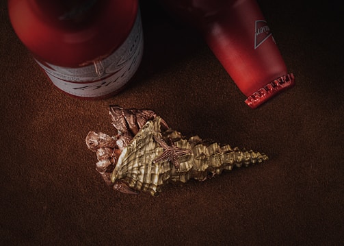 A partially unwrapped, ornate chocolate piece shaped like a horn sits alongside two red bottles on a textured brown surface. The chocolate has a gold hue with intricate patterns, while the bottles have visible labels and caps, and are positioned diagonally.