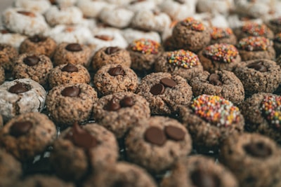 A large assortment of cookies is arranged closely together, featuring a variety of toppings such as chocolate chips, colorful sprinkles, and powdered sugar. The cookies vary in texture and appearance, indicating a diverse selection.