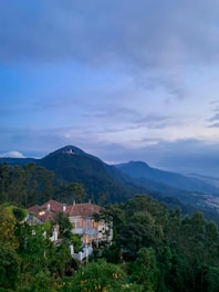 A picturesque landscape featuring a sprawling mountain range under a vast, cloudy sky. Nestled amidst lush green trees is a charming villa with large windows and a red-tiled roof. The soft evening light casts a serene glow over the scene, creating a tranquil and inviting atmosphere.