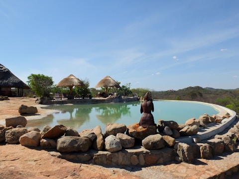 A serene landscape featuring an infinity pool surrounded by natural rock formations. In the background, there are thatched-roof structures and green foliage. A bronze sculpture of a person is positioned beside the pool, adding an artistic element. The scene is set against a backdrop of distant hills under a clear blue sky.