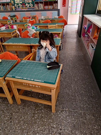 A young child wearing a face mask and a denim jacket is seated at a wooden school desk in a classroom. The classroom has several desks with orange chair covers, some of which have jackets draped over them. Bookshelves filled with books and decorations are along the back wall. The child is making a peace sign with their hand.