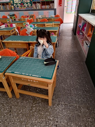 A young child wearing a face mask and a denim jacket is seated at a wooden school desk in a classroom. The classroom has several desks with orange chair covers, some of which have jackets draped over them. Bookshelves filled with books and decorations are along the back wall. The child is making a peace sign with their hand.