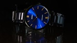 A wristwatch with a shiny blue dial featuring minimalist yellow hour markers and slim hands. The brand name is prominently displayed. The metal strap and bezel add a sleek and polished look, with the watch's reflection visible on the glossy surface below. The overall setting is dark, highlighting the vibrant blue dial.