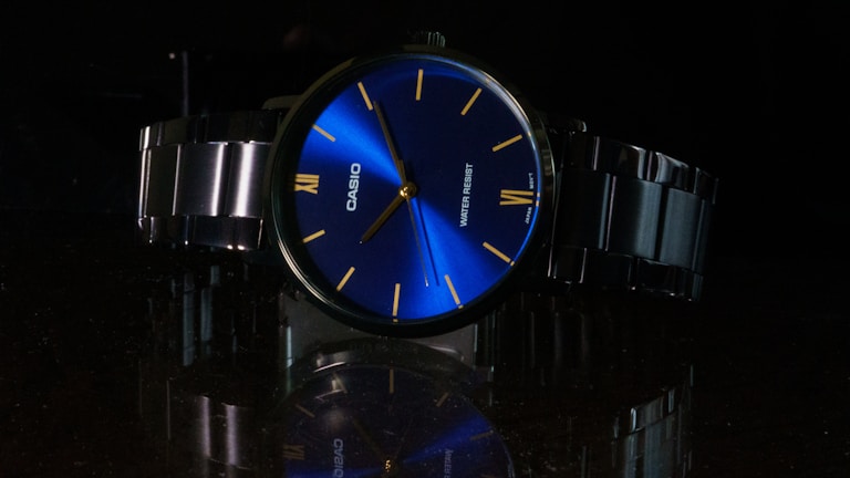 A wristwatch with a shiny blue dial featuring minimalist yellow hour markers and slim hands. The brand name is prominently displayed. The metal strap and bezel add a sleek and polished look, with the watch's reflection visible on the glossy surface below. The overall setting is dark, highlighting the vibrant blue dial.