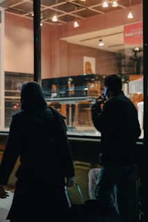 A dimly lit store window displays a grand piano. Two people, silhouetted against the light, walk by on the street outside. One is holding a phone to their ear, and the other is pulling a suitcase.