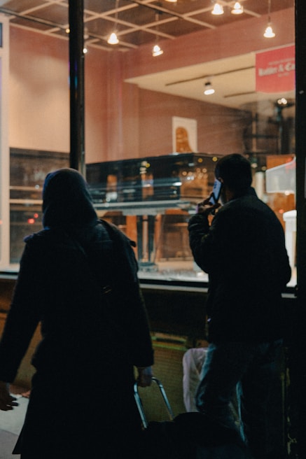 A dimly lit store window displays a grand piano. Two people, silhouetted against the light, walk by on the street outside. One is holding a phone to their ear, and the other is pulling a suitcase.