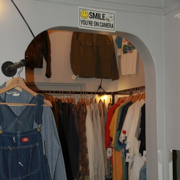 A small clothing store with a variety of garments hung on racks, showcasing coats, shirts, and denim overalls. A sign with a smiley face and the words 'Smile You're on Camera' is displayed above the entrance. The clothes are organized by type and color, creating a visually appealing arrangement.