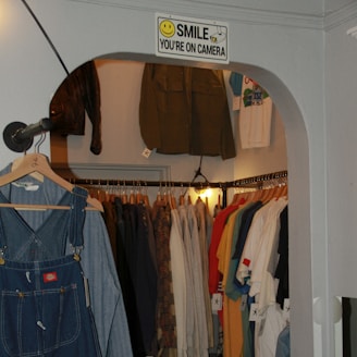 A small clothing store with a variety of garments hung on racks, showcasing coats, shirts, and denim overalls. A sign with a smiley face and the words 'Smile You're on Camera' is displayed above the entrance. The clothes are organized by type and color, creating a visually appealing arrangement.