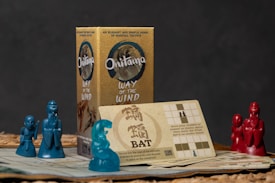 A tabletop game setup with a box labeled 'Onitama: Way of the Wind' featuring martial tactics-themed imagery. Two sets of martial figure miniatures, one in blue and the other in red, are placed on game board sheets featuring grid patterns. A card with instructions and illustrations is also prominently displayed.