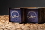 Two boxed sets of the game 'Onitama' are placed side by side on a woven surface. The boxes have a predominantly blue design with the game's name printed in white. The background is dark, highlighting the boxes.
