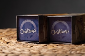 Two boxed sets of the game 'Onitama' are placed side by side on a woven surface. The boxes have a predominantly blue design with the game's name printed in white. The background is dark, highlighting the boxes.