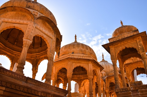Intricately carved stone pavilions with arches and domes stand against a clear blue sky, showcasing architectural details typical of Rajasthani style.