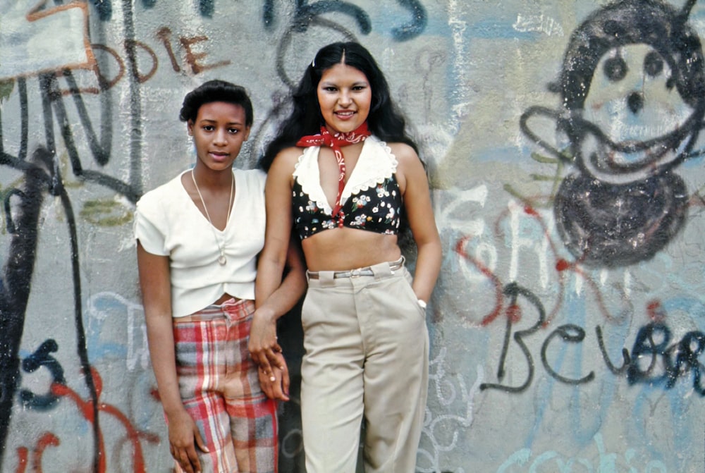 two women standing next to each other in front of graffiti