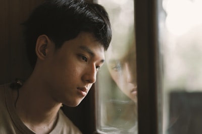 A young person gazes thoughtfully out of a window, their reflection visible in the glass. The soft lighting creates a serene, contemplative atmosphere.
