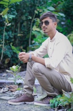 A person is squatting in a natural setting, surrounded by lush greenery. They are wearing sunglasses, a light-colored shirt, beige pants, and brown sneakers, and appear relaxed and contemplative.