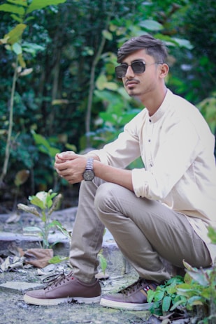 A person is squatting in a natural setting, surrounded by lush greenery. They are wearing sunglasses, a light-colored shirt, beige pants, and brown sneakers, and appear relaxed and contemplative.