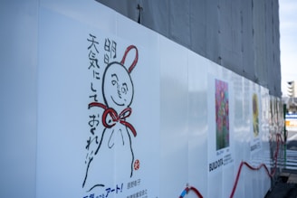 A wall with a whimsical illustration of a character featuring a round head, a smiling face, and a red ribbon tied around its neck. Japanese text accompanies the illustration. The wall also displays colorful art posters, including floral designs, with some construction materials and fencing visible.