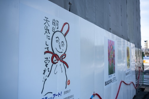 A wall with a whimsical illustration of a character featuring a round head, a smiling face, and a red ribbon tied around its neck. Japanese text accompanies the illustration. The wall also displays colorful art posters, including floral designs, with some construction materials and fencing visible.