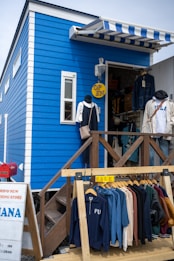 A small blue clothing store with a striped awning above the entrance. The store has an 'OPEN' sign hanging and a display rack with various jackets and hoodies at the front. The entrance is accessed via wooden stairs with a railing.