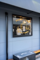 A storefront window displaying a graphic with the words 'Trunk Room Factory' and an illustration of a suitcase. Inside, a person is visible, wearing a white shirt and working in a space filled with equipment and supplies. The exterior wall is a muted blue color and there is an outdoor bench with a box featuring multiple printed logos and the word 'Hello'.