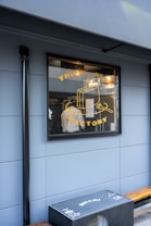 A storefront window displaying a graphic with the words 'Trunk Room Factory' and an illustration of a suitcase. Inside, a person is visible, wearing a white shirt and working in a space filled with equipment and supplies. The exterior wall is a muted blue color and there is an outdoor bench with a box featuring multiple printed logos and the word 'Hello'.