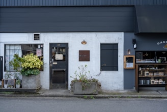 A storefront with a modern, minimalist design featuring a combination of dark grey and white walls. There are two planters with lush green plants on either side of the entrance. The front door is centered, painted black, and has a window above it with mesh-like grating. A small, discreet sign hangs above the door, showcasing the address. To the right, there is a display window with shelves containing products, possibly part of a bakery or small shop. Outdoor lighting fixtures add to the cozy yet contemporary feel.