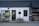 A storefront with a modern, minimalist design featuring a combination of dark grey and white walls. There are two planters with lush green plants on either side of the entrance. The front door is centered, painted black, and has a window above it with mesh-like grating. A small, discreet sign hangs above the door, showcasing the address. To the right, there is a display window with shelves containing products, possibly part of a bakery or small shop. Outdoor lighting fixtures add to the cozy yet contemporary feel.