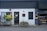 A storefront with a modern, minimalist design featuring a combination of dark grey and white walls. There are two planters with lush green plants on either side of the entrance. The front door is centered, painted black, and has a window above it with mesh-like grating. A small, discreet sign hangs above the door, showcasing the address. To the right, there is a display window with shelves containing products, possibly part of a bakery or small shop. Outdoor lighting fixtures add to the cozy yet contemporary feel.