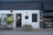 A storefront with a modern, minimalist design featuring a combination of dark grey and white walls. There are two planters with lush green plants on either side of the entrance. The front door is centered, painted black, and has a window above it with mesh-like grating. A small, discreet sign hangs above the door, showcasing the address. To the right, there is a display window with shelves containing products, possibly part of a bakery or small shop. Outdoor lighting fixtures add to the cozy yet contemporary feel.