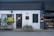 A storefront with a modern, minimalist design featuring a combination of dark grey and white walls. There are two planters with lush green plants on either side of the entrance. The front door is centered, painted black, and has a window above it with mesh-like grating. A small, discreet sign hangs above the door, showcasing the address. To the right, there is a display window with shelves containing products, possibly part of a bakery or small shop. Outdoor lighting fixtures add to the cozy yet contemporary feel.