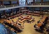 A spacious library interior with multiple long tables arranged in a square pattern, occupied by numerous people who appear to be studying or reading. The library features high ceilings, and bookshelves are visible along the upper level. The overall atmosphere suggests a focused academic environment.